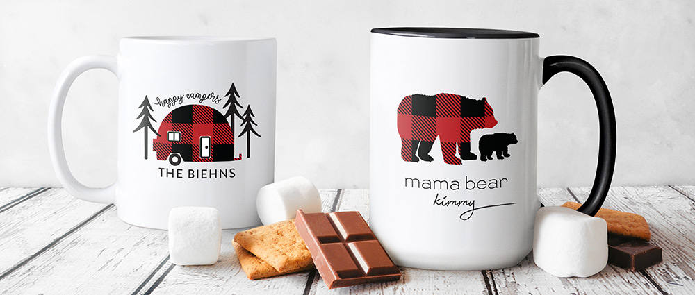 A pair of white mugs featuring custom camping designs are shown with s'more fixings
