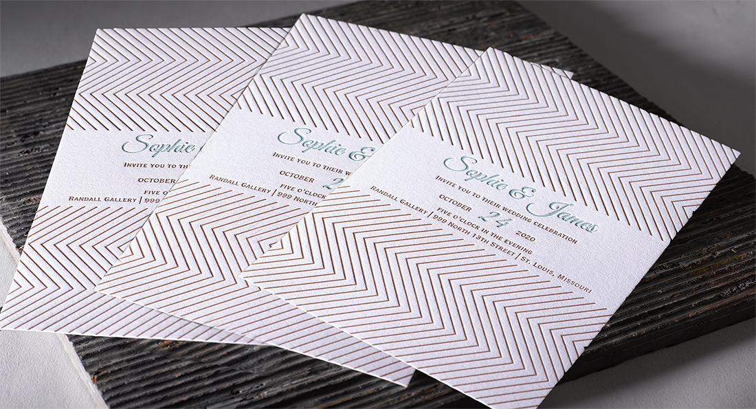 Three letterpress wedding invitations with herringbone design fanned out on a slate tile. 