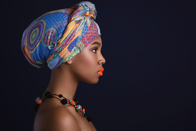A black woman standing proud with a colorful head piece, necklace and bright red lips to match. 