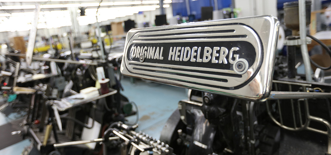 An original Heidelberg metal place is shown with more Heidelbergs in the background on a production floor.