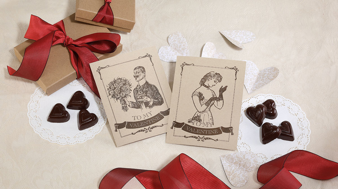 Victorian style valentines on kraft paper with brown thermography print displayed with gift boxes, red ribbon and chocolates. 