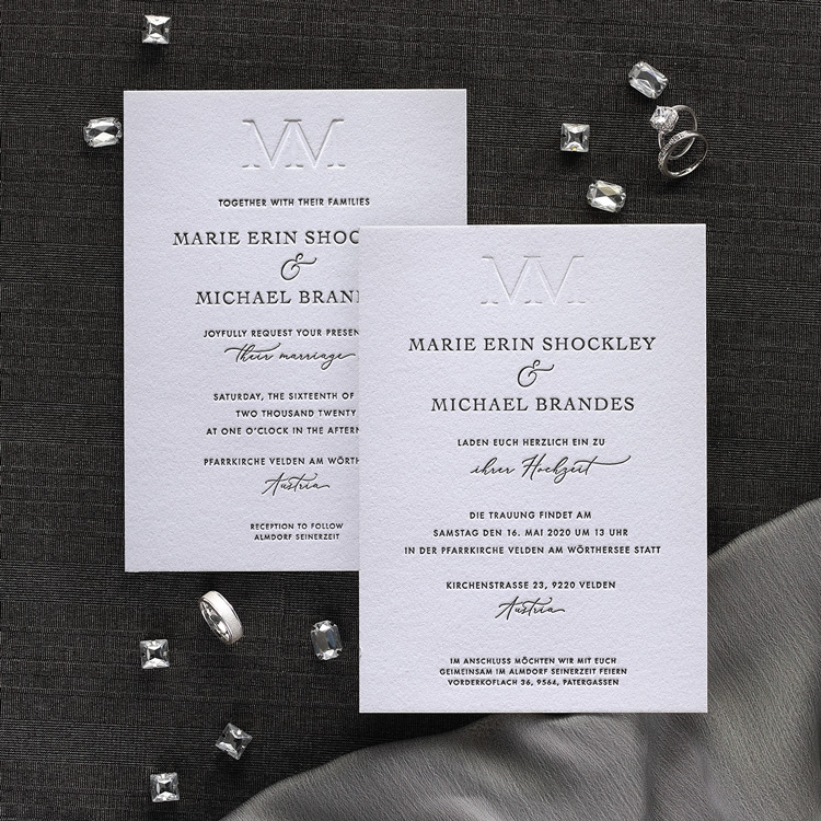 An elegant wedding invitation with blind embossed monogram at the top is shown with two rings and diamonds on a gray background. 