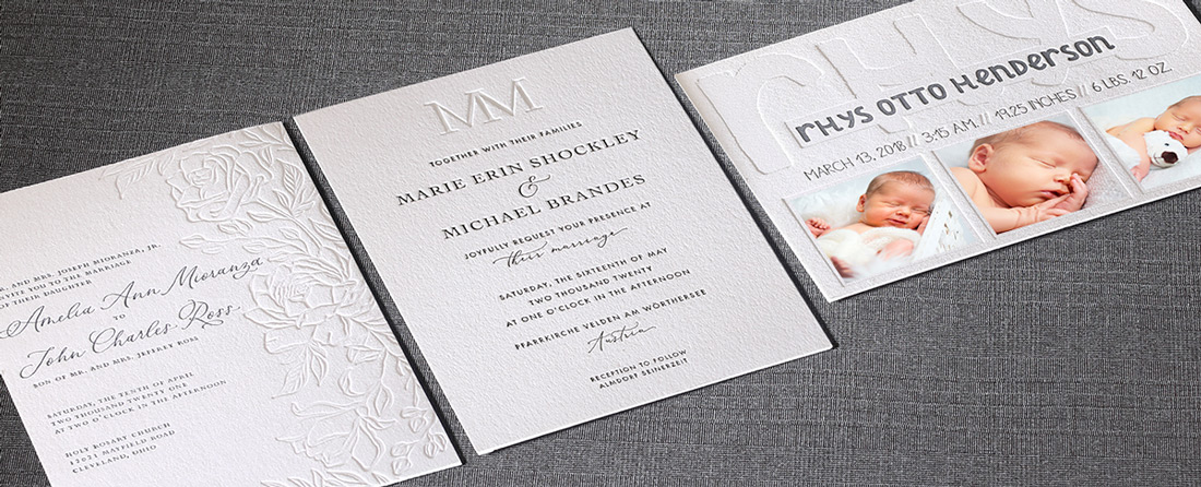 Blind embossed wedding invitations and a birth announcements are laid next to each on a gray fabric background. 
