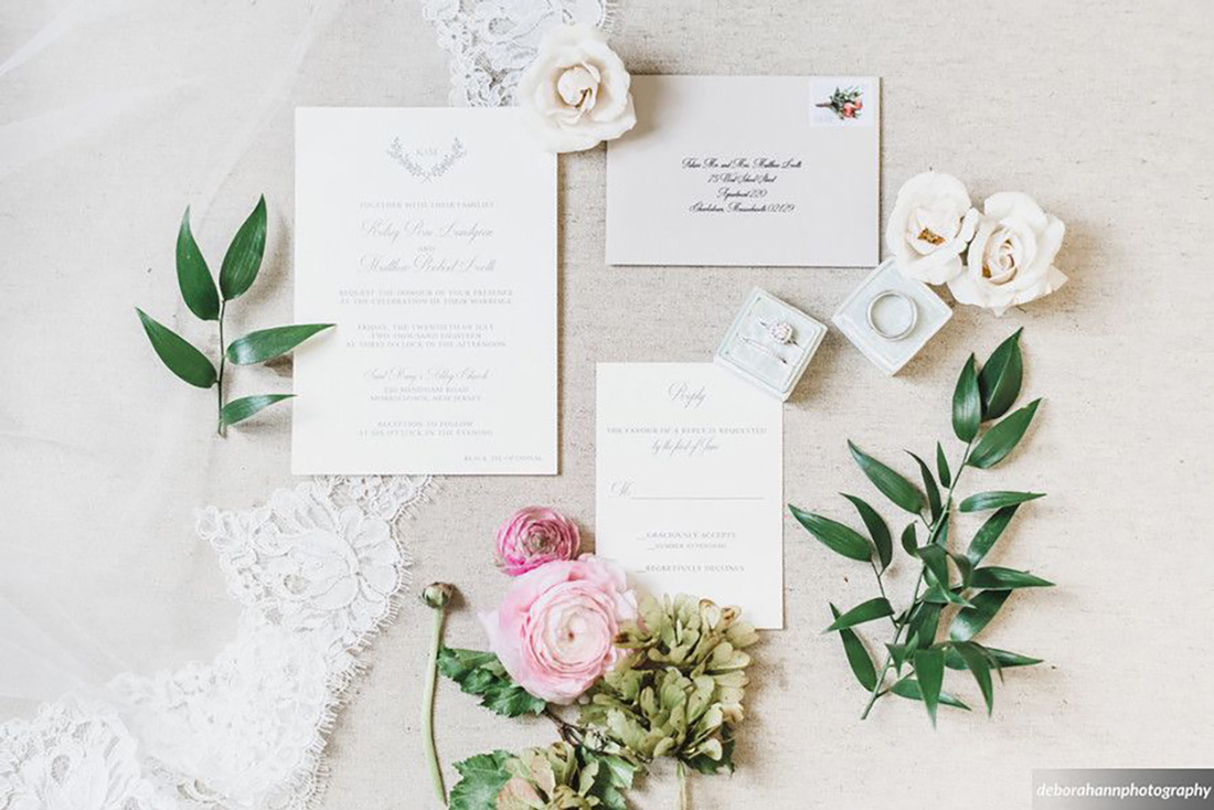 A custom wedding invitation suite by designer, Jan Halbert, is shown with floral and greenery props as well as a lace accent. 