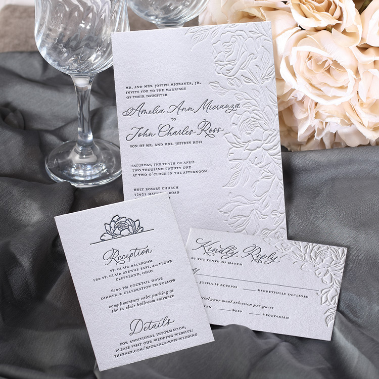 A floral wedding invitation featuring blind letterpress is shown with coordinating pieces against a gray satin backdrop and white roses. 
