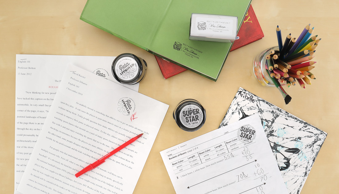 A table is shown with custom teacher stamps being used on pages of homework next to books and colored pencils. 
