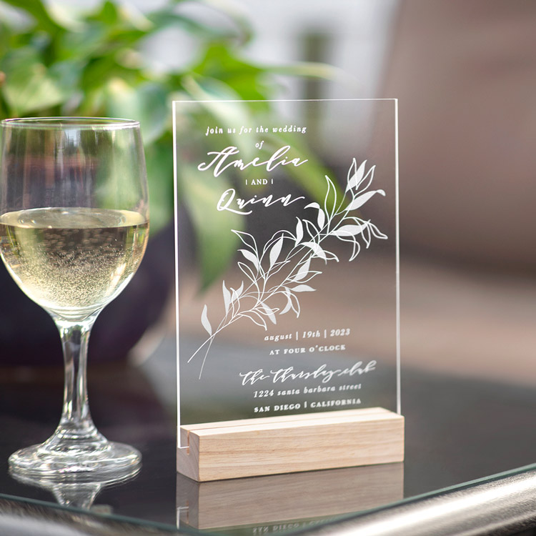 A custom acrylic wedding invitation is shown displayed on a reception table next to a glass of wine. 