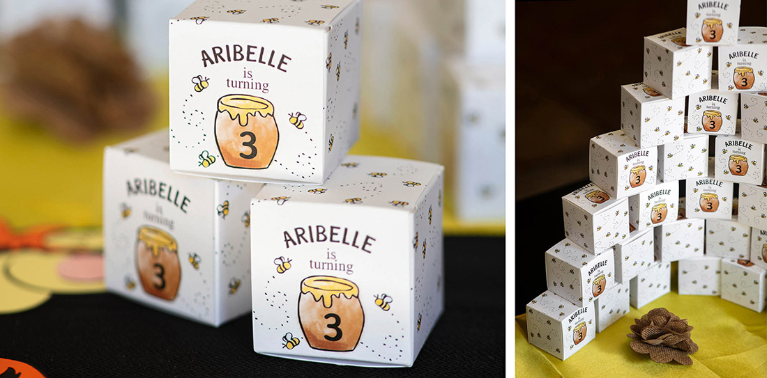 Custom favor boxes featuring a honey pot with the number three are stacked in a tower.