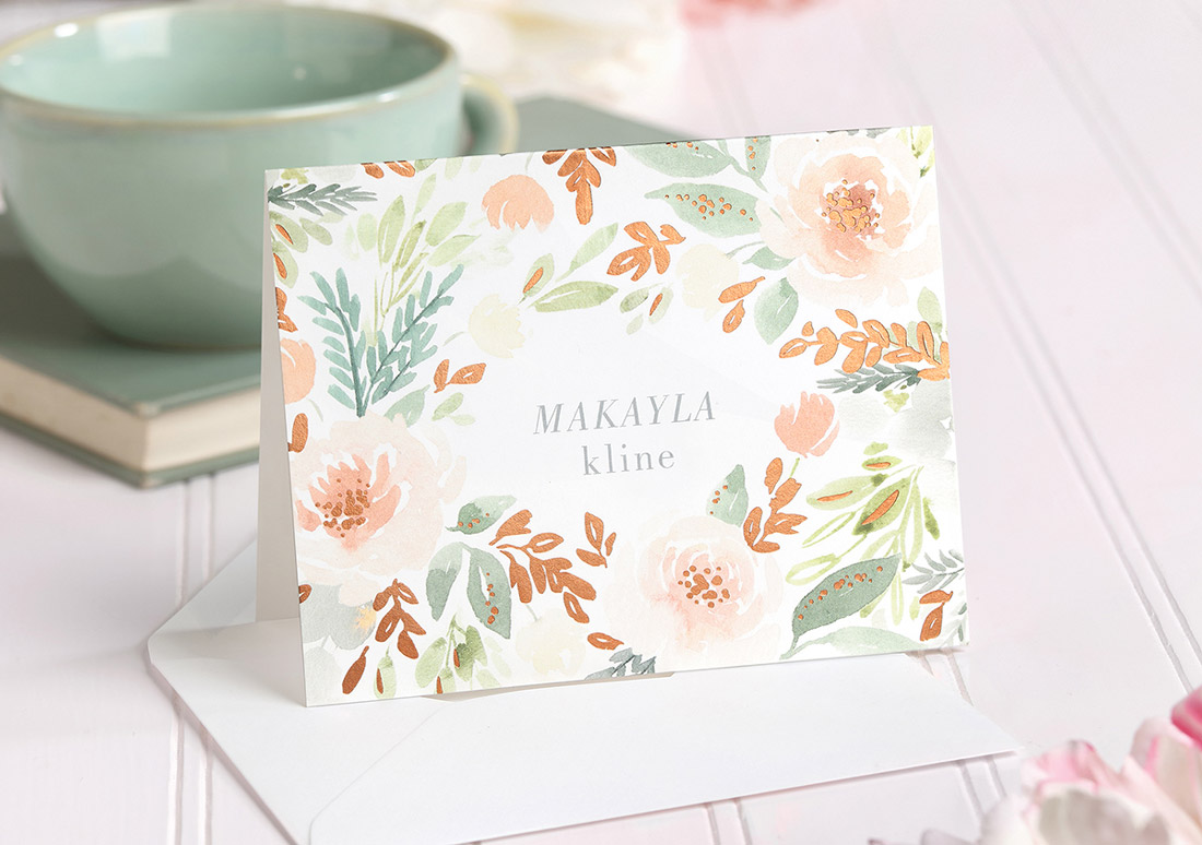An elegant folding note card with floral artwork framing a person's name is shown with a tea cup in the background. 