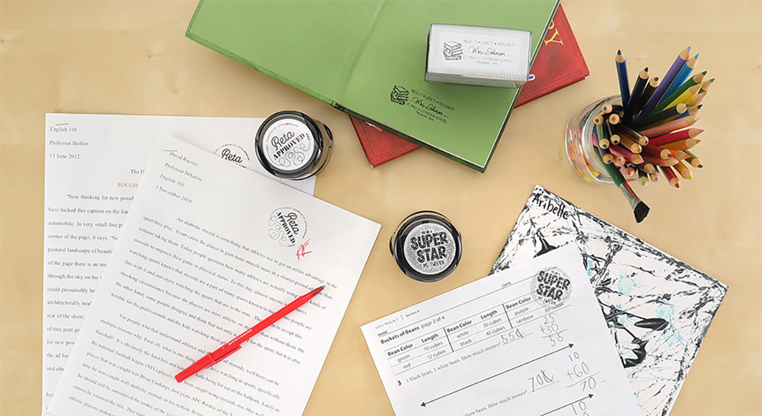 A teacher's desk with students' homework is shown with custom stamps featuring positive messages. 