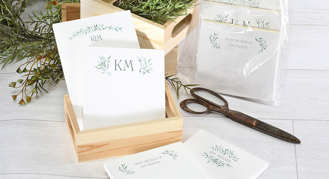 A set of custom personalized stationery is shown in a wooden display case with greenery in the background.