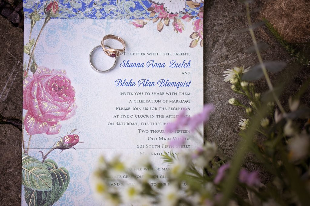 An image of the writer's wedding invitation with colorful floral design and wedding rings stacked on top. 